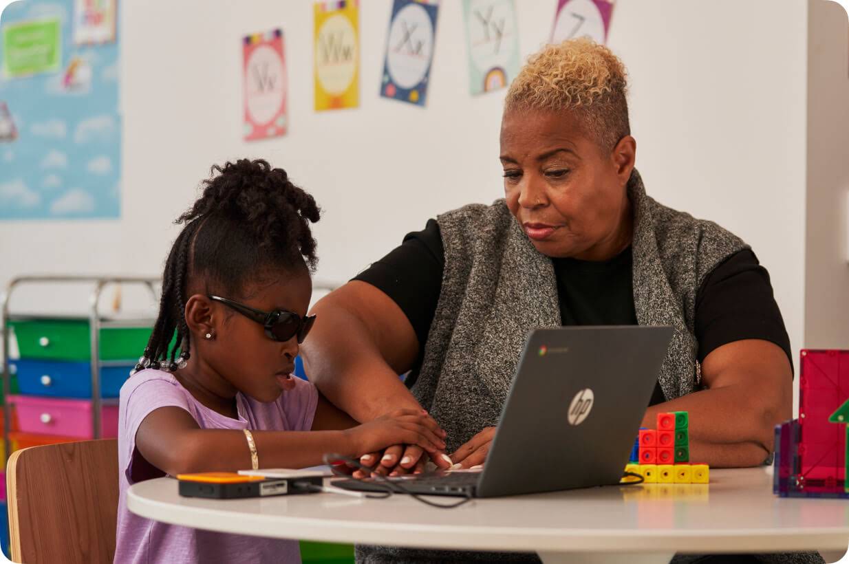 A young, visually-impaired student uses a Chromebook with the help of a braille reader while their teacher, sitting next to them, guides their hand.