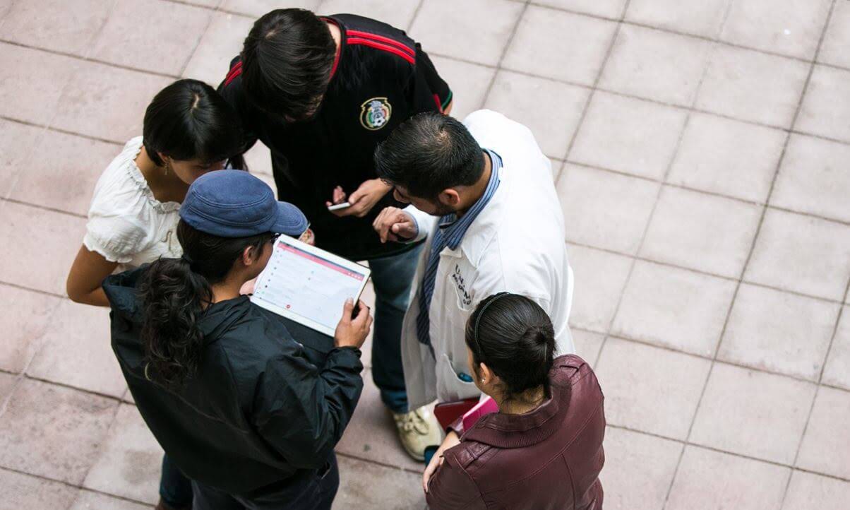 A group of students in the quad gather around as a professor leans in to review a project in Google Workspace.