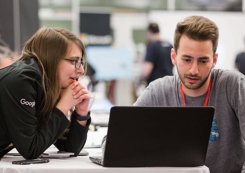 Two college aged individuals, possibly older, look at a Chromebook screen. One is wearing a Google jacket.