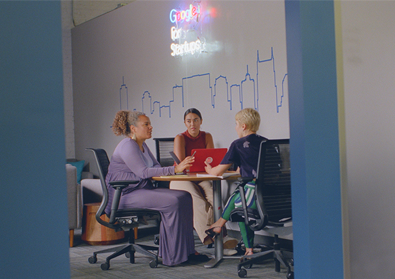 three women sitting around a table with a laptop talking.